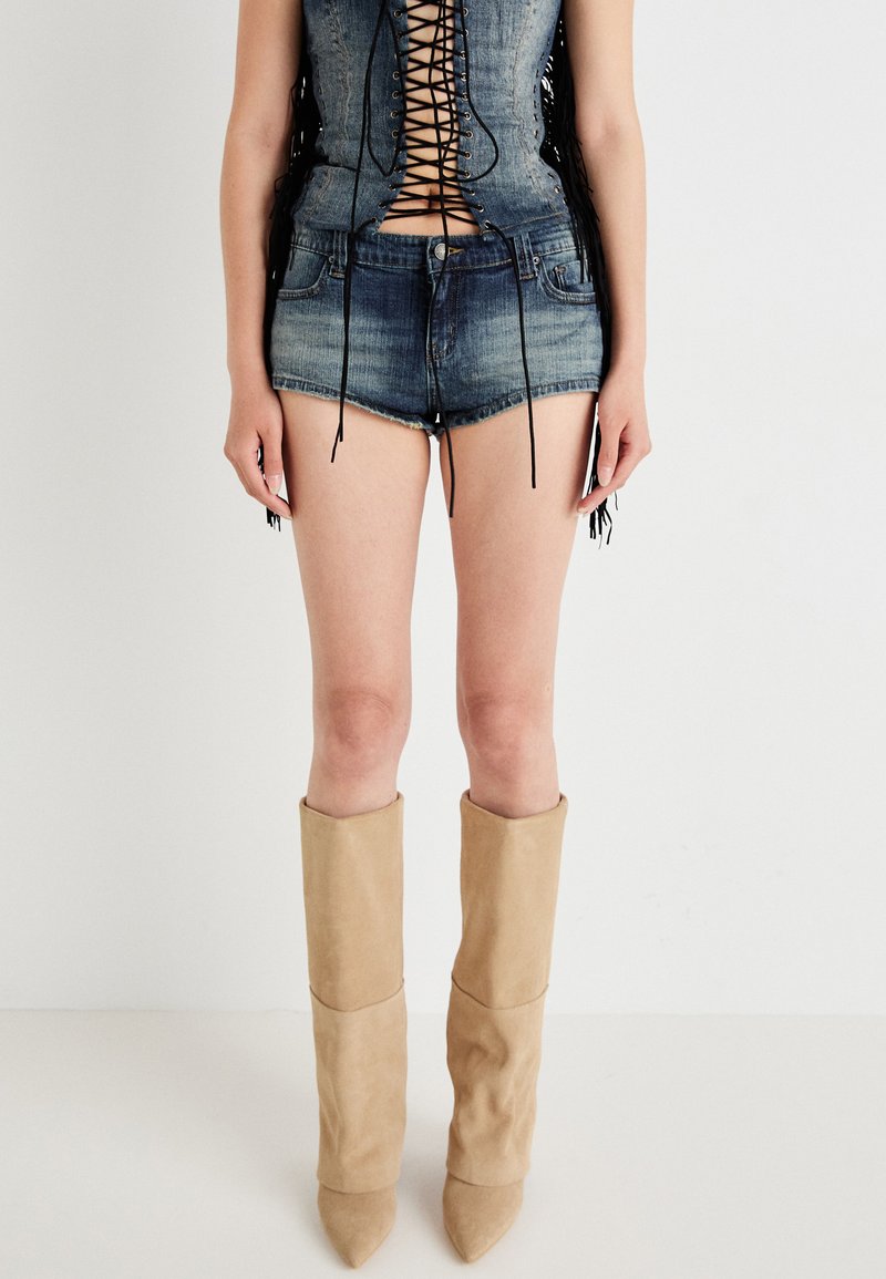 Modell i en snörd denimtop och denimshorts, bärande höga beige stövlar. Bakgrunden är helt vit, vilket fokuserar uppmärksamheten på outfiten.