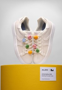 White leather sneakers with colorful plastic charms including flowers and smiley faces, displayed on a textured yellow pedestal.