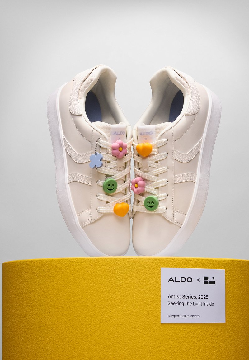 White leather sneakers with colorful plastic charms including flowers and smiley faces, displayed on a textured yellow pedestal.