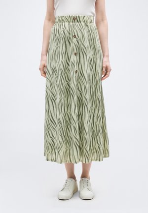 Kobieta ubrana w zieloną spódnicę midi w wzory z guzikami z przodu, białą koszulkę na ramiączkach i białe trampki, stojąca na jednolitym tle.