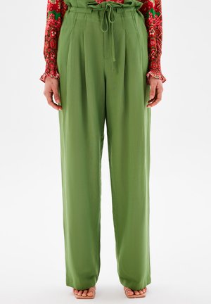 Grüne, lockere Hose mit Kordelzugbund, Falten und einer glatten Textur, kombiniert mit offenen Schuhen.