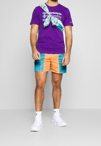 Fialové tričko s logem Columbia, doplněné oranžovými a tyrkysovými vzorovanými šortkami. Bílé tenisky a vzorovaný doplněk přehozený přes rameno.