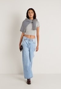 Γκρι κοντομάνικο πουκάμισο, ανοιχτό μπλε τζιν με φαρδύ πόδι, γκρι πλεκτό φορεμένο γύρω από τους ώμους, σκουρό καφέ μποτάκια μέχρι τον αστράγαλο, κρατώντας μαύρη τσάντα φάκελο.