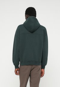 Haut à capuche vert en tissu doux. Présente une capuche, des poignets et un ourlet côtelés, avec une coupe décontractée et des coutures sur le dos.