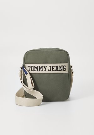 Sac bandoulière vert olive avec une large sangle beige et une bande avec le texte « Tommy Jeans » sur le devant, sur un fond clair uni.