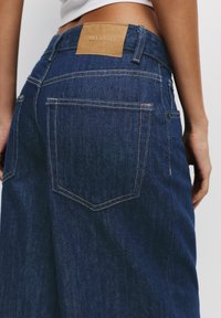 Jean en denim bleu foncé avec une taille mi-haute, doté d'un patch en cuir beige portant l'inscription "PULL&BEAR" et de poches arrière standard avec une couture contrastante.