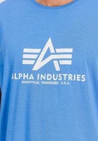 Modré bavlněné tričko s bílou grafikou znázorňující hvězdu a křídla, na kterém je zobrazen název značky "ALPHA INDUSTRIES" spolu s textem o umístění.