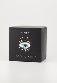 Boîte noire présentant un design stylisé d'œil avec un centre aqua, des accents dorés, et le texte "TIMEX" et "JACQUIE AICHE" en blanc.