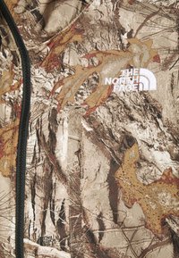 Tkanina w wzór kamuflażu w ziemistych odcieniach brązu, beżu i pomarańczu. Posiada czarny zamek błyskawiczny i białe logo 'The North Face'.