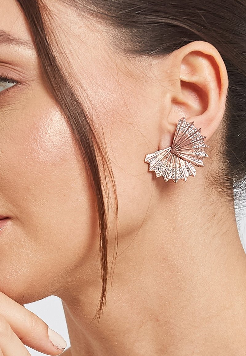 Femme portant une boucle d'oreille en forme d'éventail en or rose avec de petites pierres scintillantes, cheveux glissés derrière l'oreille, et doigts près du menton.