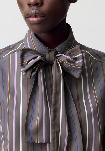 Blusa a righe nei toni del marrone, grigio e lavanda; presenta un ampio colletto a fiocco e un tessuto liscio con una leggera lucentezza.