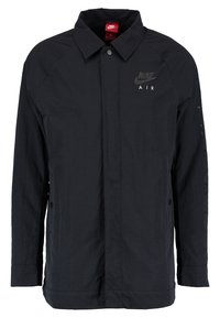 Černá lehká bunda s límečkem, zapínáním na knoflíky a logem "Nike Air". Má dlouhé rukávy a minimální strukturu.