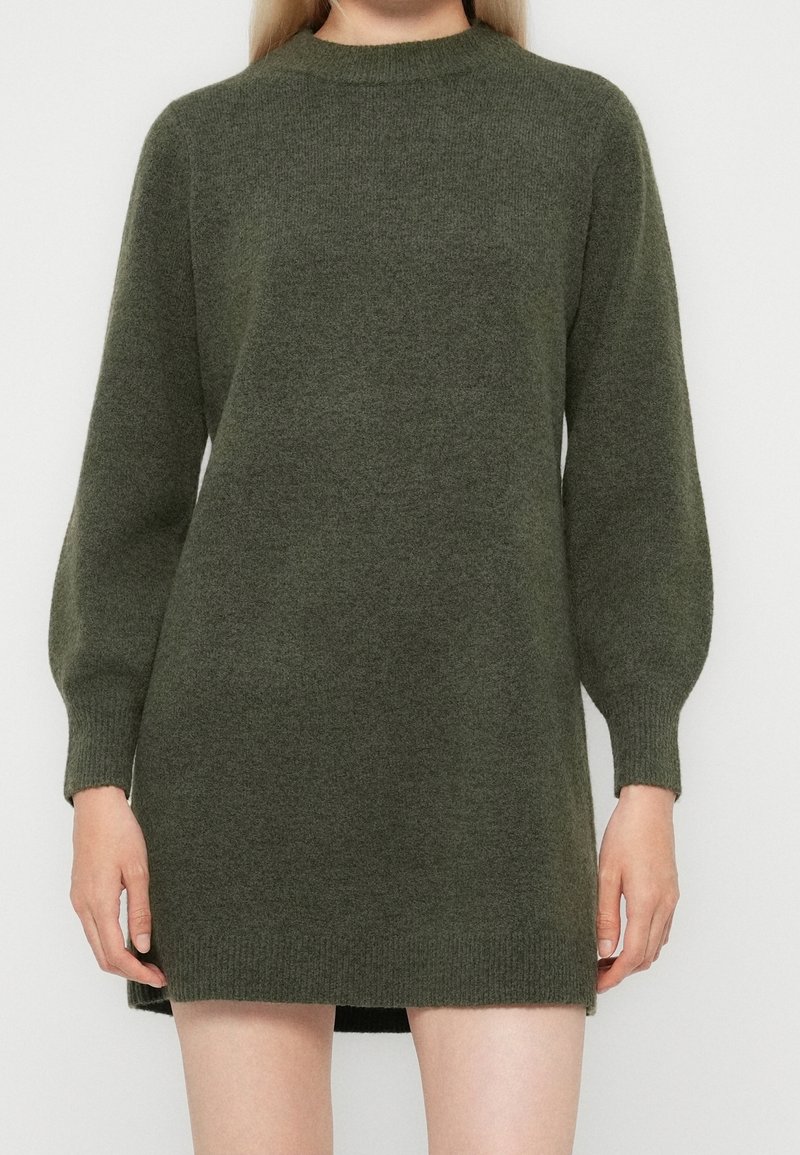 Femme portant une robe-pull tricotée à manches longues de couleur vert olive avec des poignets et un ourlet côtelés, debout devant un fond uni.