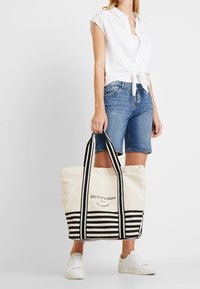 Plátěná taška v krémové barvě s horizontálními černými pruhy, se silnými černými uchy a potiskem loga umístěným ve středu přední strany.