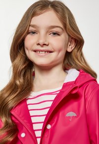 Ryški rožinis vandeniui atsparus lietpaltis su kapišonu, su sidabrinio skėčio logotipu. Po juo matomas baltos juostelės marškinėliai.