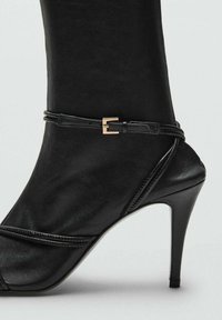 Talons aiguilles noirs avec un bout pointu, fabriqués en cuir lisse, présentant une sangle croisée avec une boucle en ton doré.