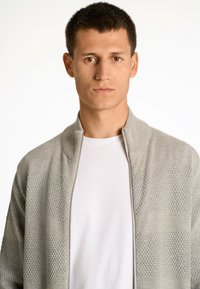 Grau melierte Zip-Top-Strickjacke mit strukturierten Paneelen und glatten Abschnitten, getragen über einem weißen Hemd. Der Stoff wirkt weich und leicht.