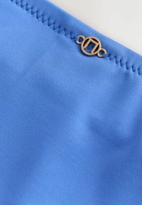 Modrá tkanina s hladkým povrchom, s pozláteným kruhovým kovovým detailom a prešitým okrajom.