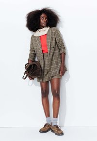 Blazer en tartan marron avec des shorts assortis, top rouge, chaussettes grises et chaussures à motif léopard marron. Porte un sac en cuir texturé marron.