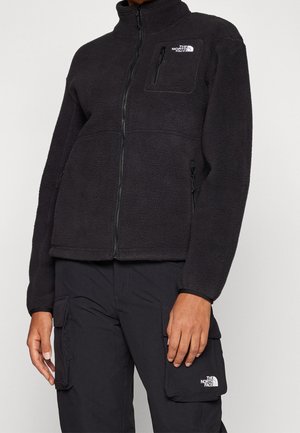 Osoba ubrana w czarną kurtkę polarową i spodnie typu cargo, oba z logo The North Face, stojąca na tle jednolitego jasnego tła.