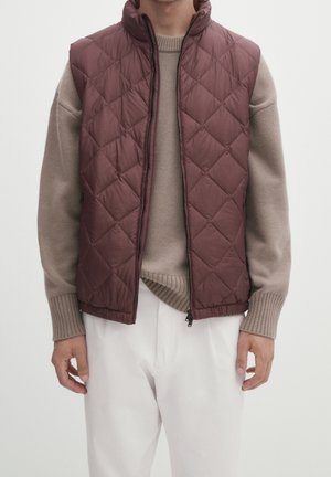 Osoba ubrana w brązową pikowaną kamizelkę bez rękawów na beżowym dzianinowym swetrze oraz białe spodnie na jednolitym tle.