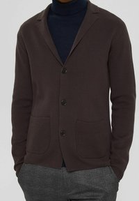 Brauner Cardigan mit strukturiertem Strickdesign, zwei vorderen Taschen, einem Reverskragen und schwarzen Knöpfen, getragen über einem navyblauen Rollkragenpullover.