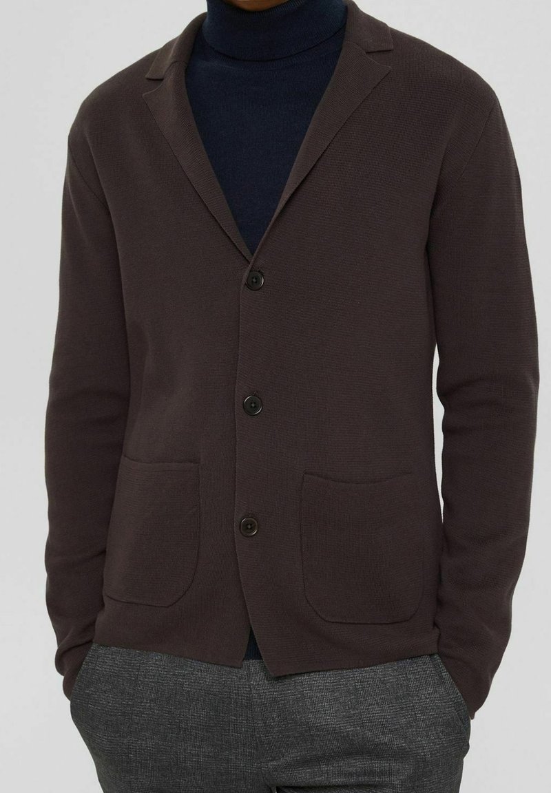 Brauner Cardigan mit strukturiertem Strickdesign, zwei vorderen Taschen, einem Reverskragen und schwarzen Knöpfen, getragen über einem navyblauen Rollkragenpullover.