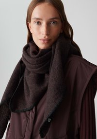 Frau mit heller Haut und langen braunen Haaren, die einen dunkelbraunen Schal um ihren Hals gewickelt und eine passende braune Weste über einem gerippten Pullover trägt.