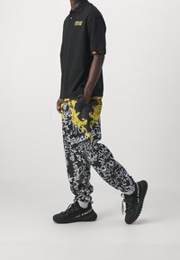 Czarna koszulka polo z żółtym logo, zestawiona z czarnymi spodniami z białym graffiti i żółtymi ornamentami. Czarne sneakersy z logo.