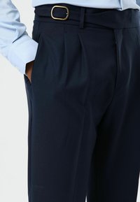 Marineblauwe plooirok met gestructureerde stof, voorzien van zijzakken en een riem met een rechthoekige messing gesp. Gestyled met een lichtblauw overhemd.