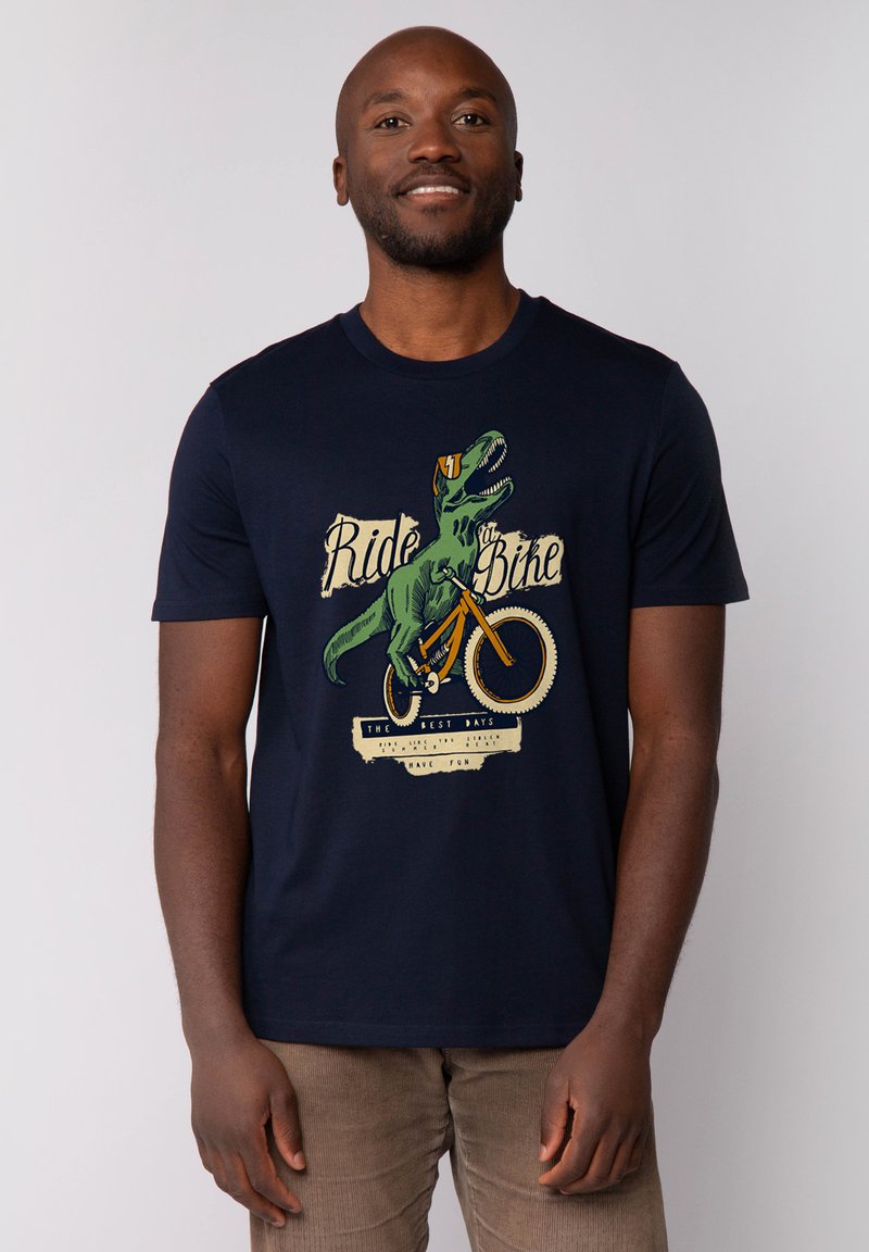 Marineblauwe t-shirt van katoen, met een grafische print van een dinosaurus die op een fiets rijdt, met tekst eronder, wat een leuk thema illustreert.