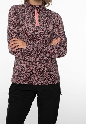 Kvinde iført pink og sort leopardprint kvart-lynlås fleece med sorte bukser, armene krydset, foran ensfarvet hvid baggrund.