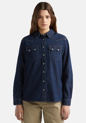 Junge Frau steht frontal, trägt ein dunkelblaues Jeanshemd mit Knöpfen und beige Hosen vor einem schlichten weißen Hintergrund.