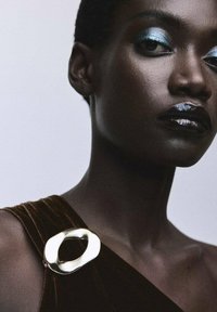 Acercamiento de un modelo que lleva una prenda de terciopelo oscuro con un gran broche ovalado de oro liso en el hombro y maquillaje destacado.