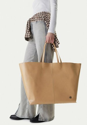 Persona con vaqueros gris claro, camiseta blanca de manga larga y camisa de cuadros atada a la cintura sostiene un gran bolso de mano de cuero marrón claro.