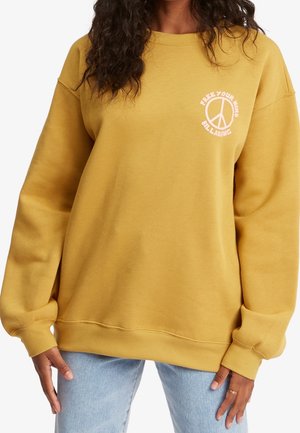 Personne portant un sweat-shirt jaune moutarde avec un signe de paix blanc et le texte « Free Your Mind Billabong », associé à un jean bleu clair.