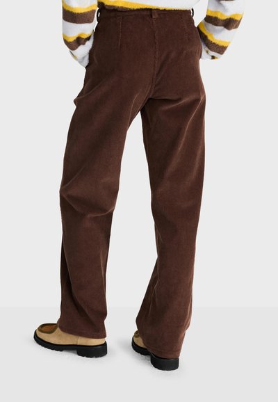 Personne portant un pantalon en velours côtelé marron ample et des chaussures beiges, debout, les mains dans les poches, vue de dos sur un fond clair.