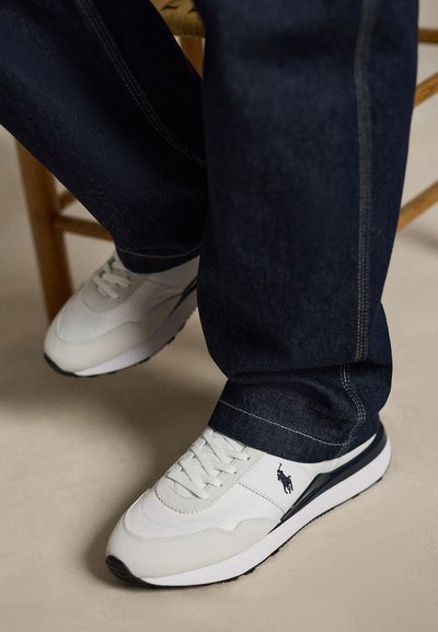 Adidași albi din piele cu logo polo negru pe lateral, purtați cu blugi albastru închis, așezați pe un podea bej lângă un picior de scaun din lemn.