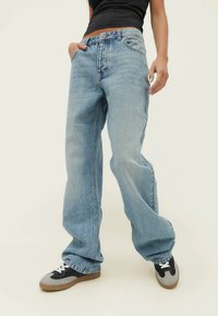 Person som bär ljusblå vida jeans och svarta och grå sneakers med vita snören, står med ena handen i fickan.