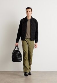 Veste aviateur noire, pantalon olive, chaussures noires, portant un sac en cuir noir avec un accent circulaire. Design minimaliste, texture lisse.
