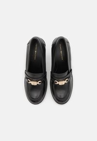 Mocasines de cuero negro con detalle de cadena dorada en la parte superior, marcados como "Tommy Hilfiger" en el interior, mostrados desde arriba sobre un fondo blanco.