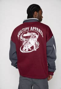 Bordeauxrotes und graues Sweatshirt mit einem großen weißen Motiv eines Hundes und einer Person auf der Rückseite. Detailliertes Design mit einem runden Rahmen und Text.