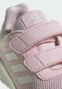 Zapatilla deportiva rosa con materiales de malla y cuero, cintas de velcro, tres rayas blancas y un patrón texturizado en la capa exterior.