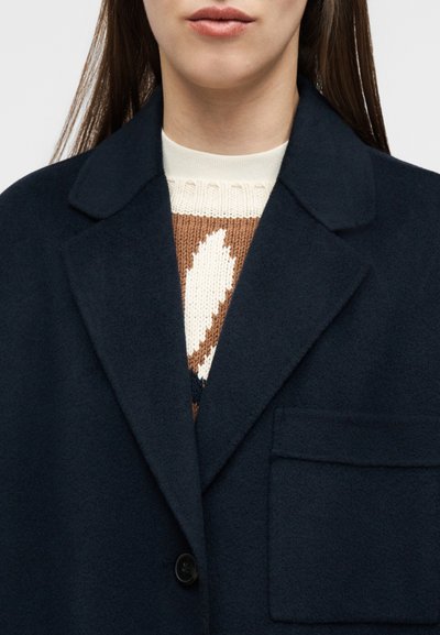 Γυναίκα που φοράει σκούρο μπλε μάλλινο παλτό με μεγάλη τσέπη και κουμπί πάνω από καφέ και κρεμ πουλόβερ με σχέδιο πλεξίματος.