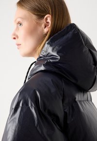 Námorníckomodrá puffrová bunda s lesklým povrchom, so šnúrkou na kapucni a mierne oversized strihom.