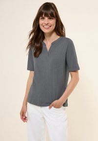 Chemise grise à manches courtes et col en V, avec une coupe décontractée et des manches retroussées. Fabriquée en matériel léger. Portée avec un pantalon blanc.