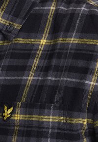 Camisa de franela en negro y gris con patrón de cuadros amarillos, con cuello abotonado y un bolsillo parche con un logo bordado.