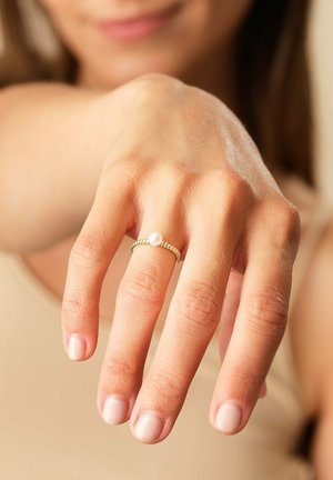 Delicate gold ring with a twisted band and central white pearl, displayed on a hand with natural nail polish against a neutral background.