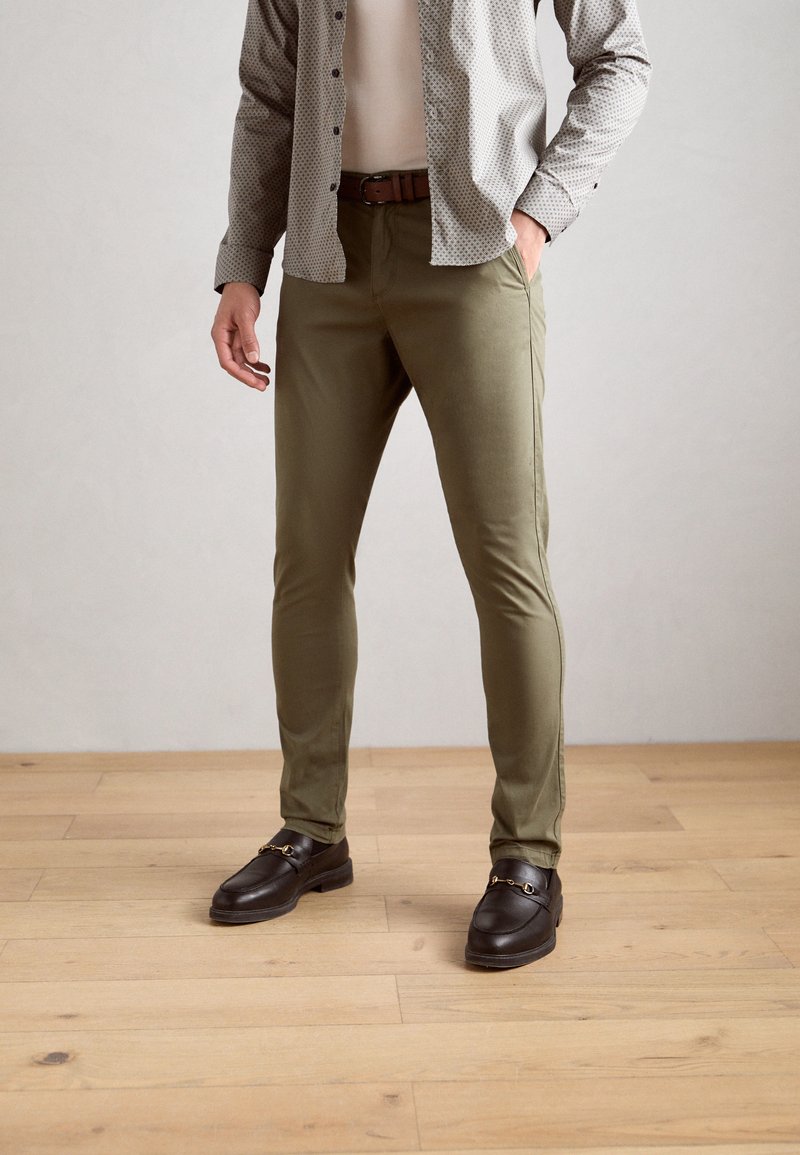 Pantalones verde oliva en corte ajustado, hechos de material suave. Combinados con una camisa gris de estampado y zapatos negros con acentos de hardware dorado.