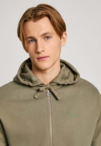 Sudadera verde oliva con cremallera y capucha ajustable, de tela texturizada y un pequeño logo verde de "Pepe Jeans" en el pecho.